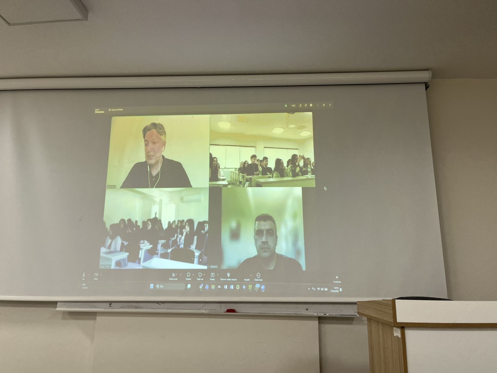 Projeksiyon perdesini yakından gösteren bu kare, çevrimiçi toplantının dijital arayüzüne odaklanıyor. Ekranda, telekonferans yoluyla bağlanan konuğunuzun ve toplantıya katılan diğer kişilerin görüntüleri yer alıyor. Aynı zamanda sınıfın ekrana yansıyan görüntüleri de mevcut, bu da söyleşinin "iki taraflı" interaktif doğasını vurguluyor.